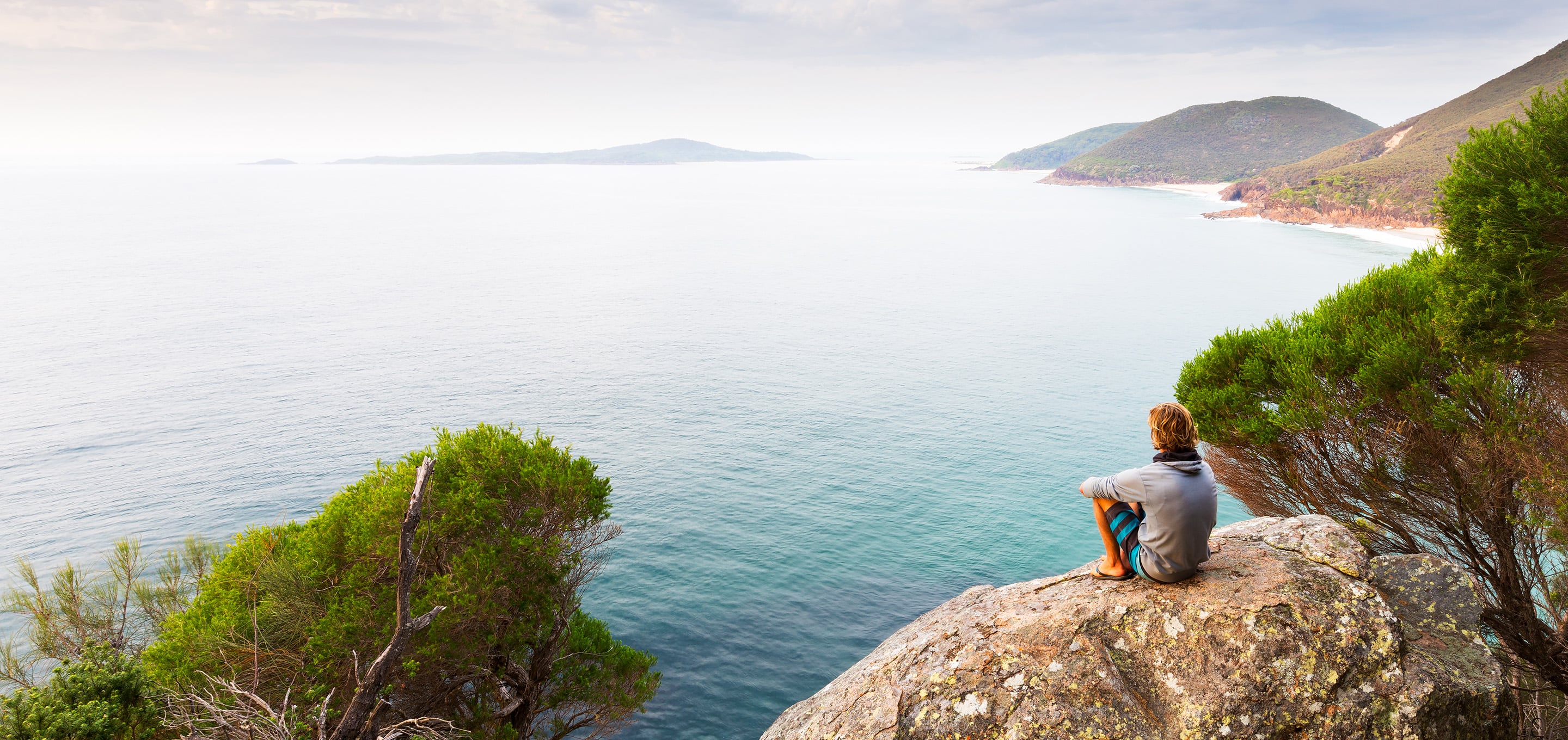 A person sitting on a rocky overlook, looking out over open ocean and coastline